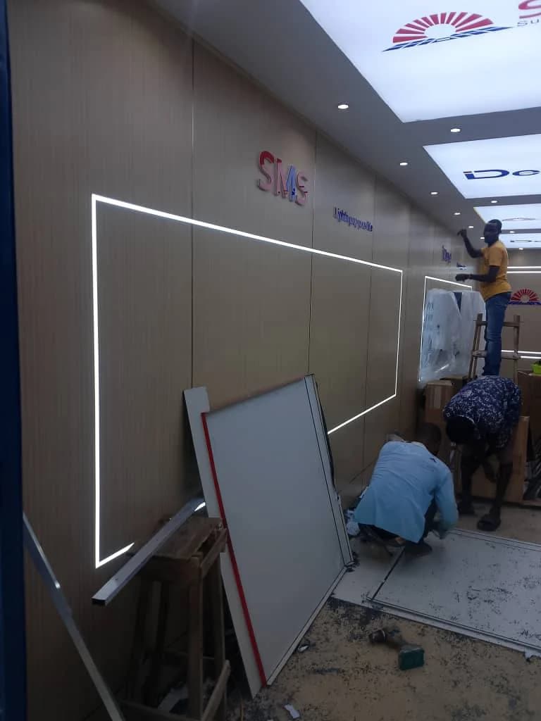 Fabrication technicians installing recessed LED linear lighting and wood-grain cladding in a SIMS retail showroom in Lagos.