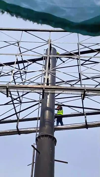 Rigger in safety harness standing on a catwalk of a tall steel unipole billboard structure against the sky.