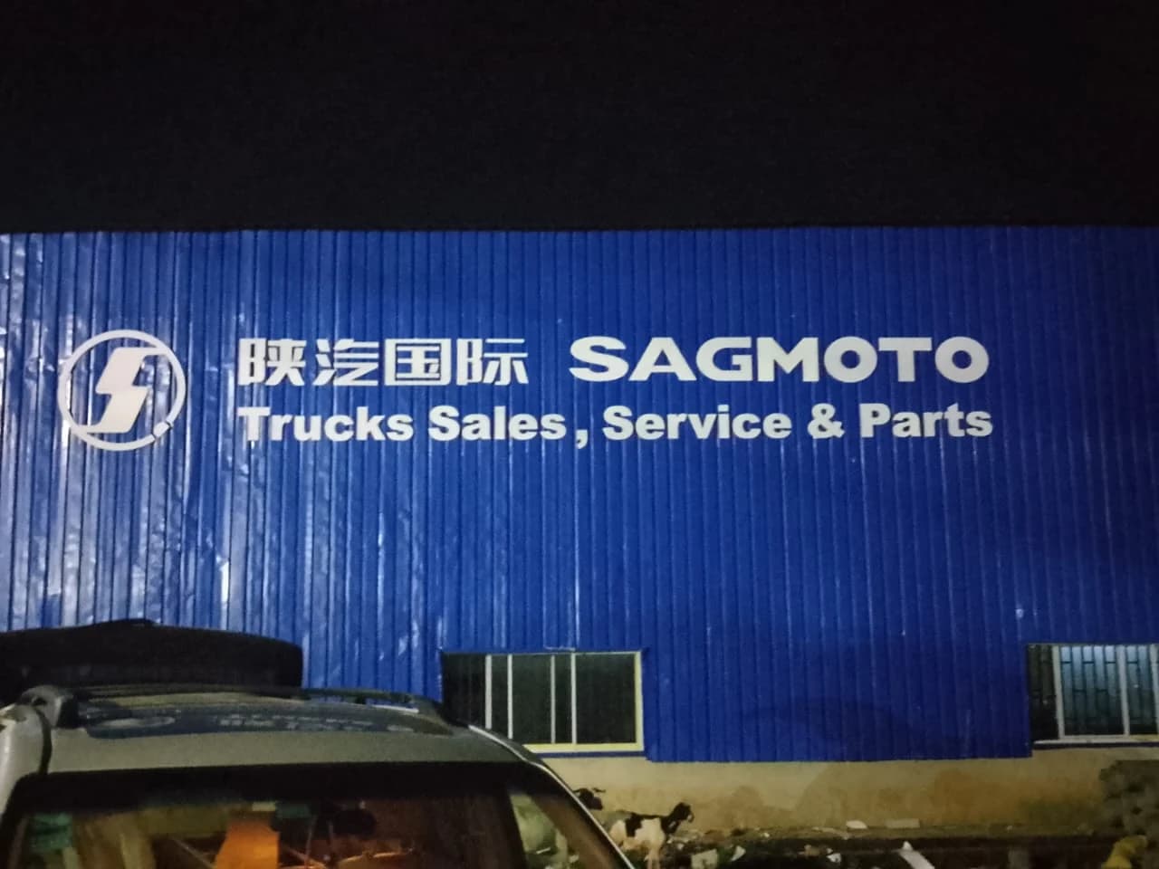 Large white SAGMOTO Trucks sign and logo emblem mounted on blue corrugated industrial warehouse cladding.
